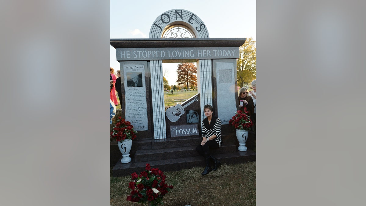 Nancy Jones in a black and white striped sweater and black pants sitting in front of a monument for George Jones
