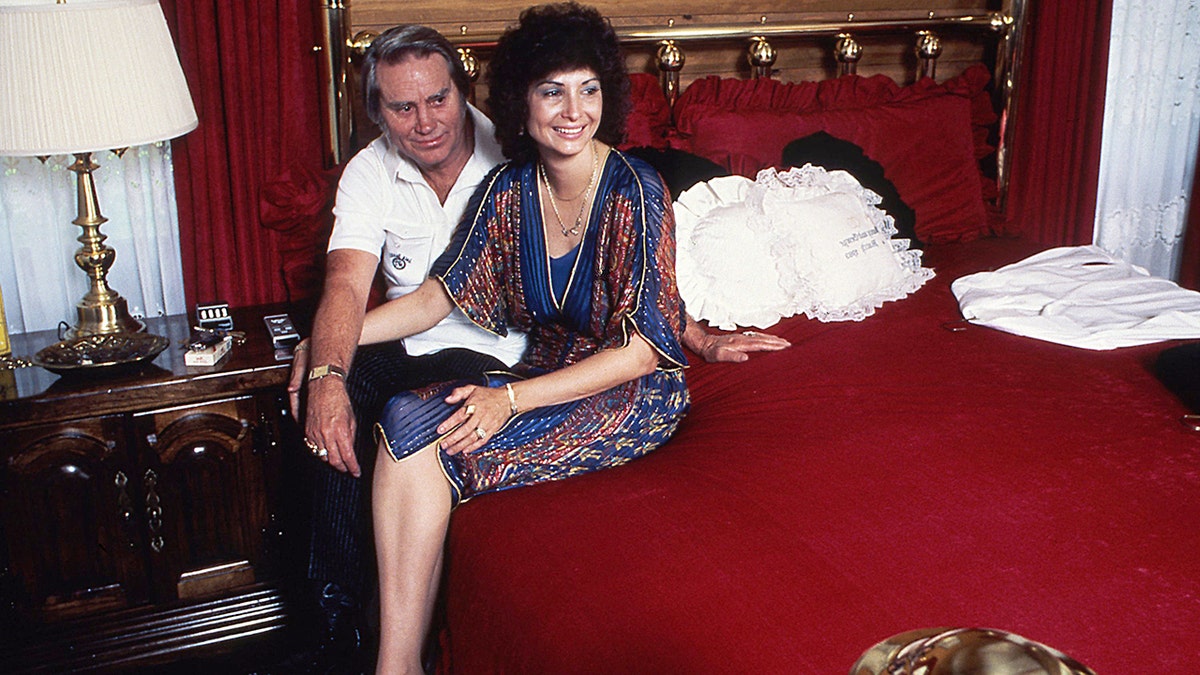 Nancy Jones in a multiprinted dress sitting next to George Jones wearing a white shirt and black pants on a red bed
