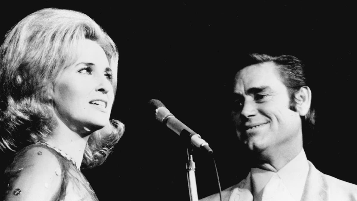 A close-up of Tammy Wynette singing iin front of a mic as George Jones smiles at her