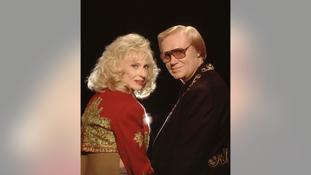 Tammy Wynette wearing a red and gold jacket next to George Jones in a black shirt and sunglasses
