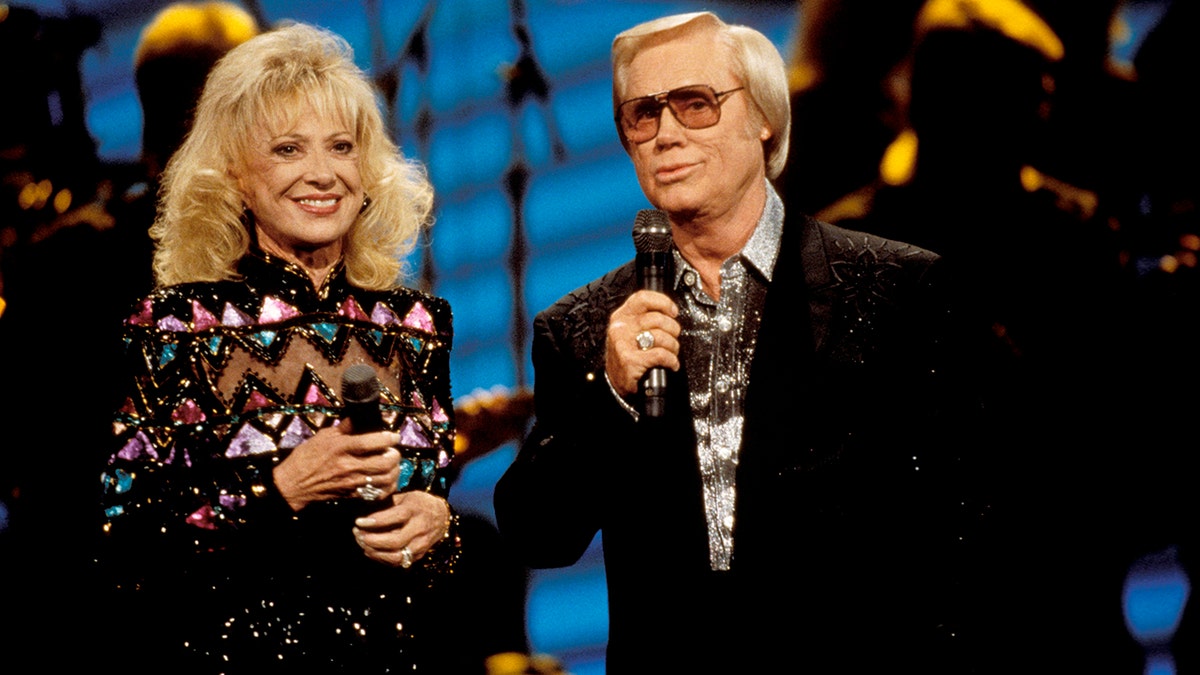 Tammy Wynette wearing a multicolored dress next to George Jones in a black suit holding a mic