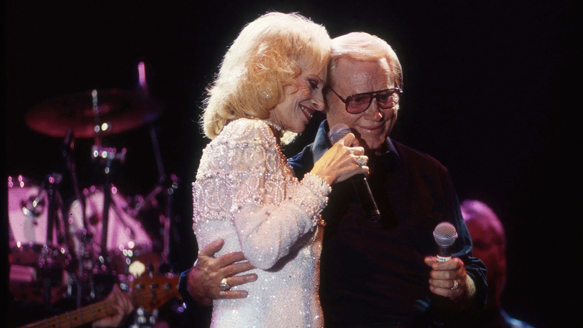 Tammy Wynette wearing a white dress being embraced by 