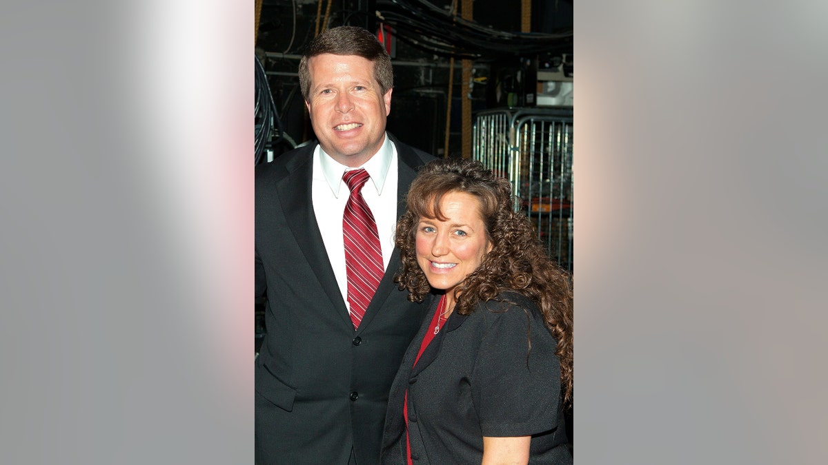 Jim Bob vestindo um terno escuro e gravata cor de vinho ao lado de sua esposa Michelle Duggar usando um vestido preto