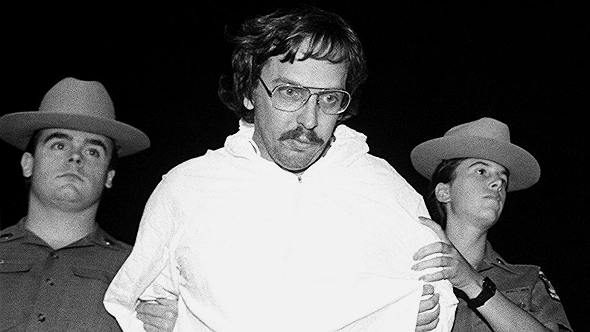 Joel Rifkin in a white sweater being held by two police officers