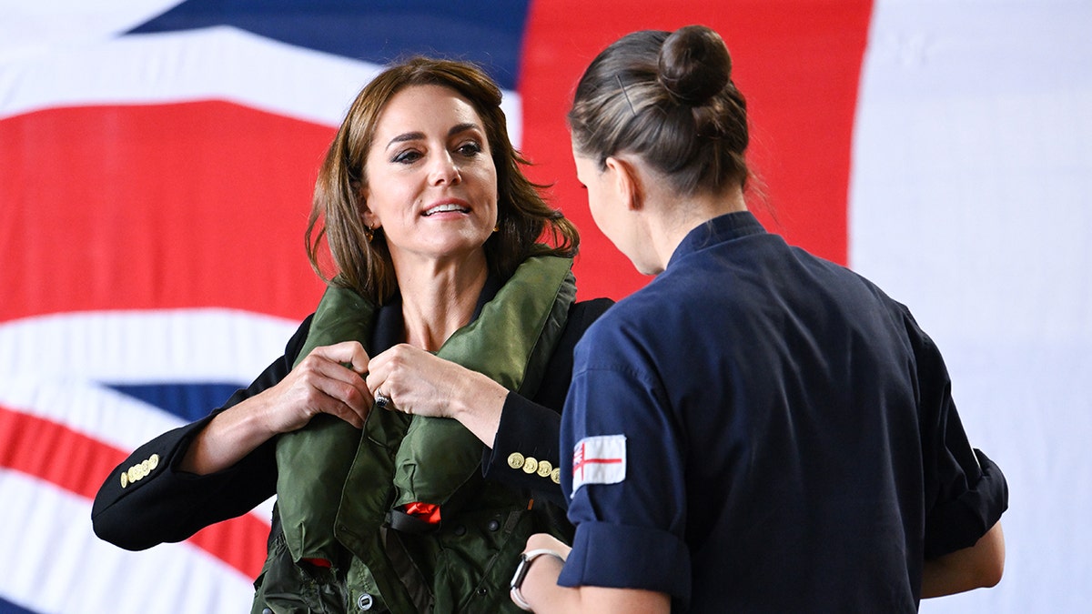 Kate Middleton wearing a hunter green jacket talking to a woman in a navy jumpsuit in fron the Union Jack