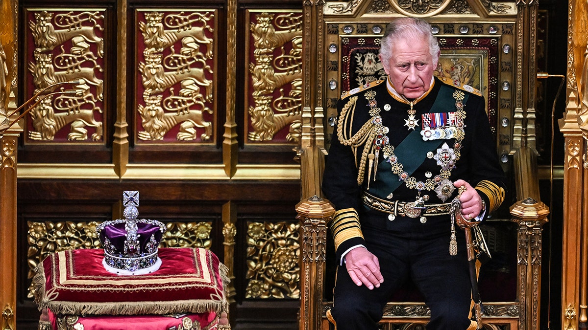 King Charles wearing a formal suit with medals sitting next to a crown