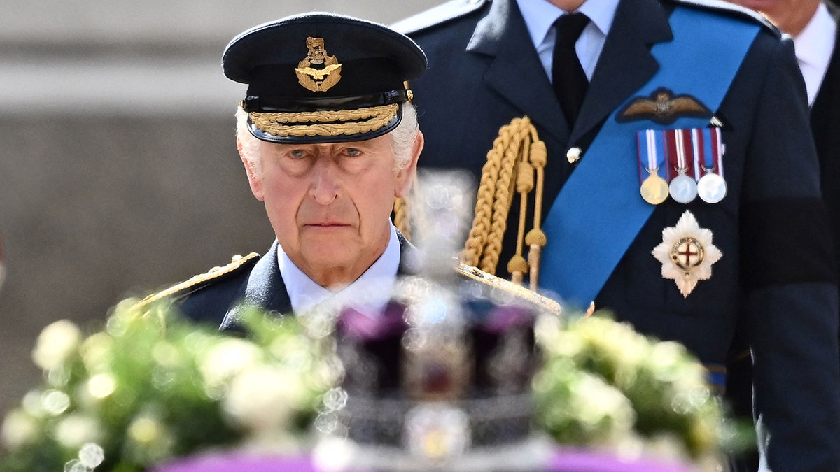 King Charles in a military suit looking sadly at his mothers coffin