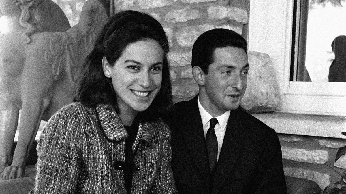 A close-up of Marina Karella smiling in a coat next to Prince Michael of Greece in a suit and tie