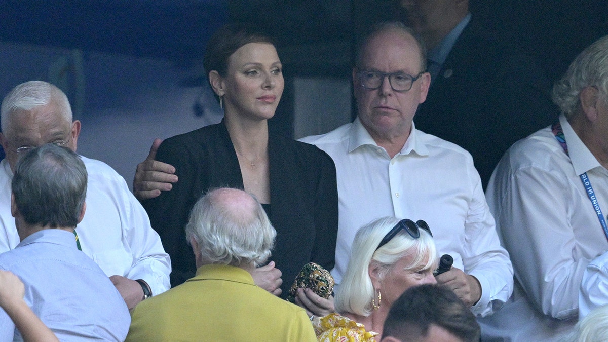 Prince Albert wearing a white shirt putting his arm around Princess Charlene in a black blazer