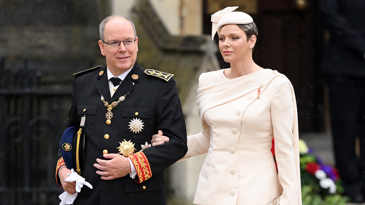 Princess Charlene wearing a pale pink dress while in the arm of Prince Albert wearing a suit with medals