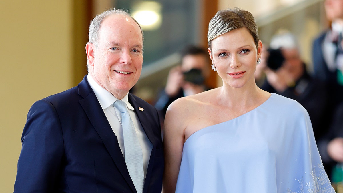 Princess Charlene wearing an off the shoulder blue dress standing next to Prince Albert in a navy suit
