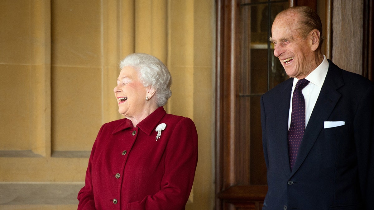 Queen Elizabeth wearing a red coat and laughing next to a smiling Prince Philip in a suit and tie