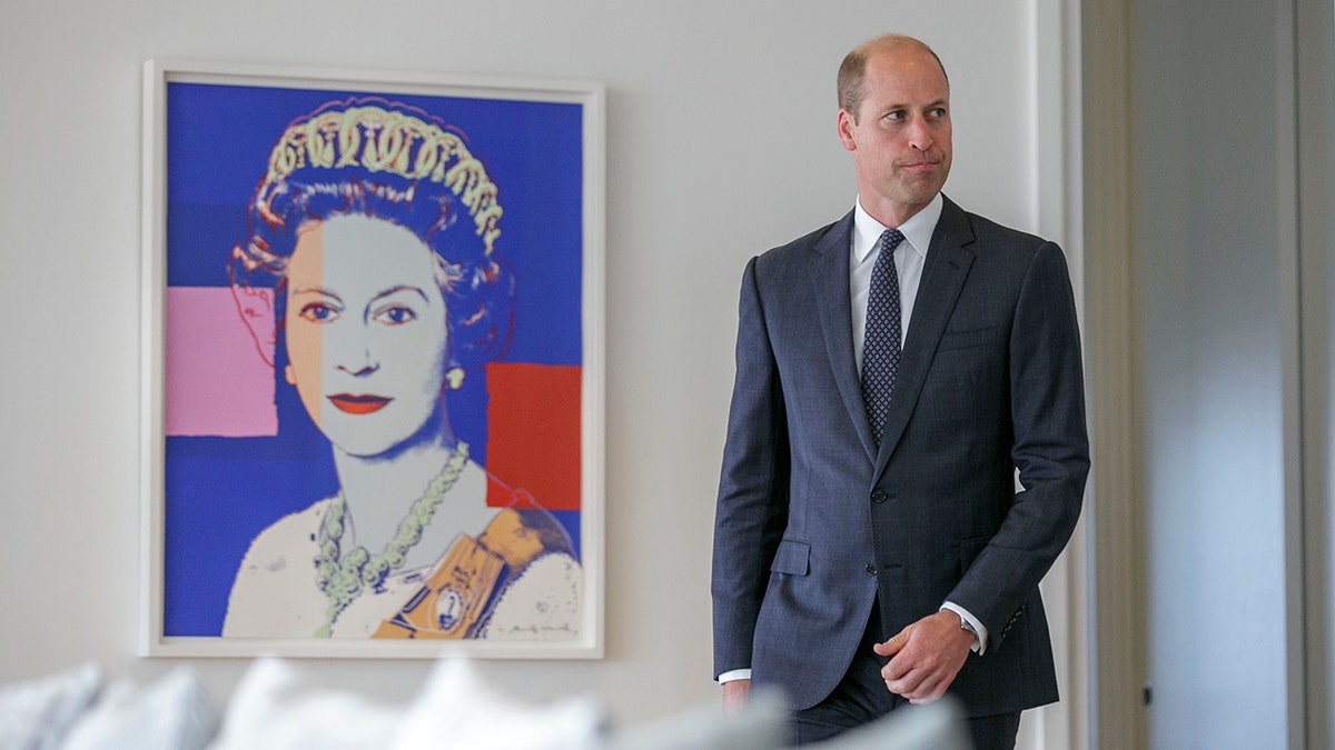 Prince William in a navy suit walking next to a Warhol portrait of his grandmother Queen Elizabeth II
