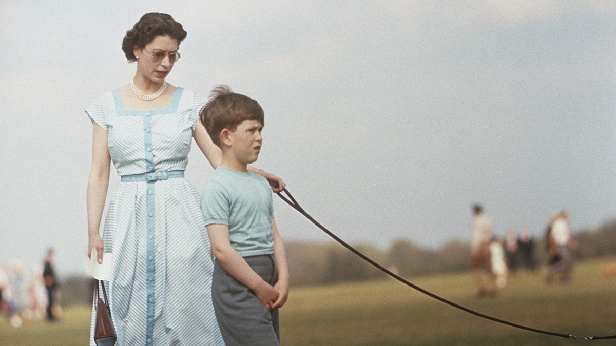 Queen Elizabeth wearing a light blue dress holding a leash next to Prince Charles in a matching shirt and grey shorts
