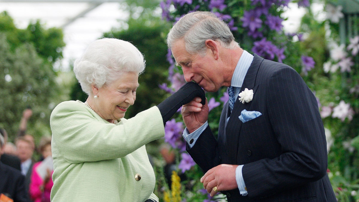 Prince Charles wearing a dark blue suit kissing his mothers hand, who is wearing a light green dress