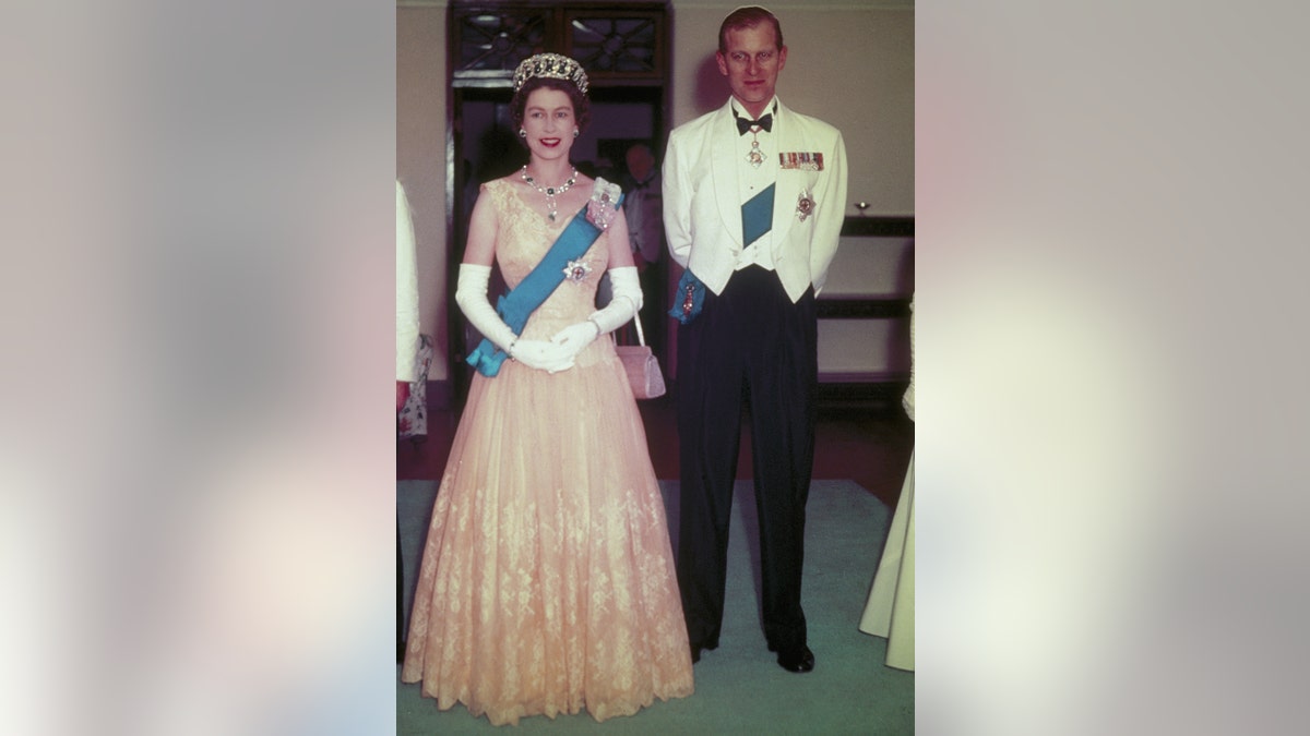 Queen Elizabeth wearing a white gown with a blue sash standing next to Prince Philip in a white tux