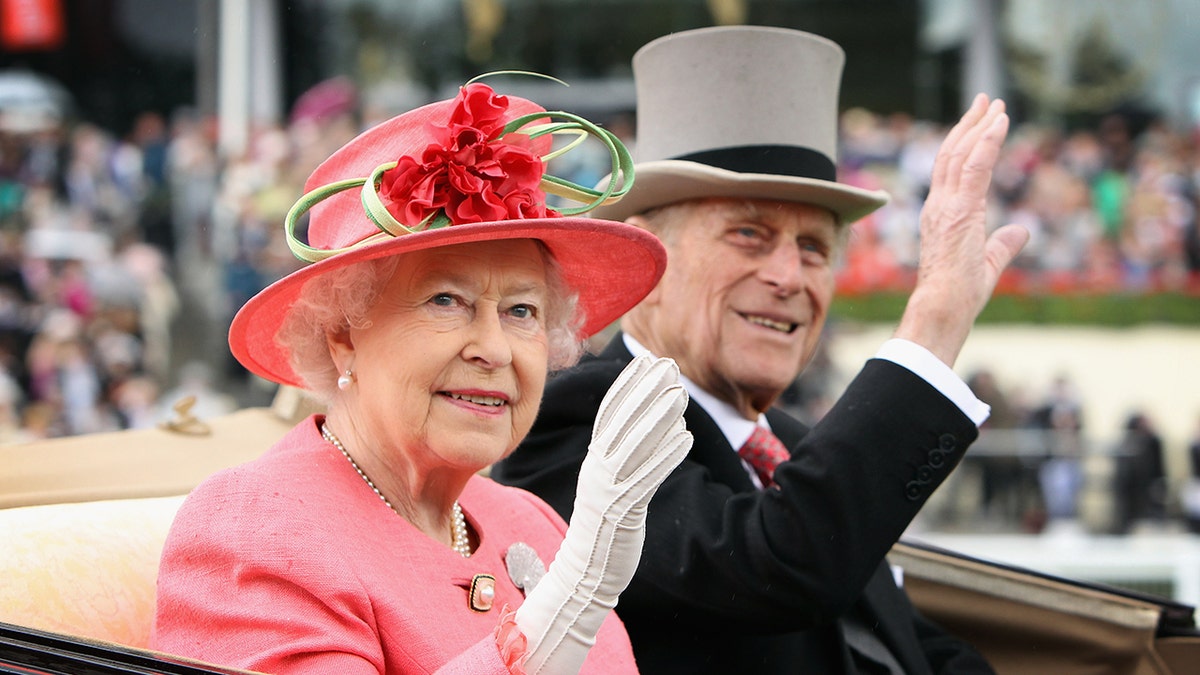 Queen Elizabeth, wearing a coral dress and matching hat, waves next to Prince Philip, who is wearing a black suit and gray top hat.