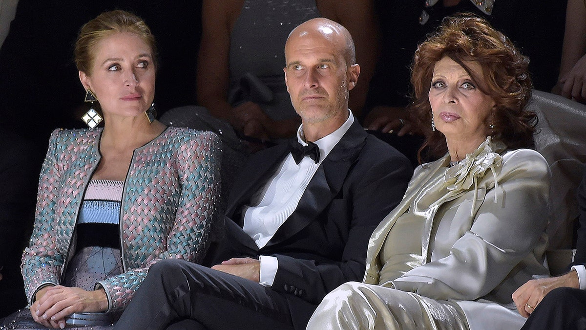 Sophia Loren in a golden suit sitting next to her son and a woman at a fashion show