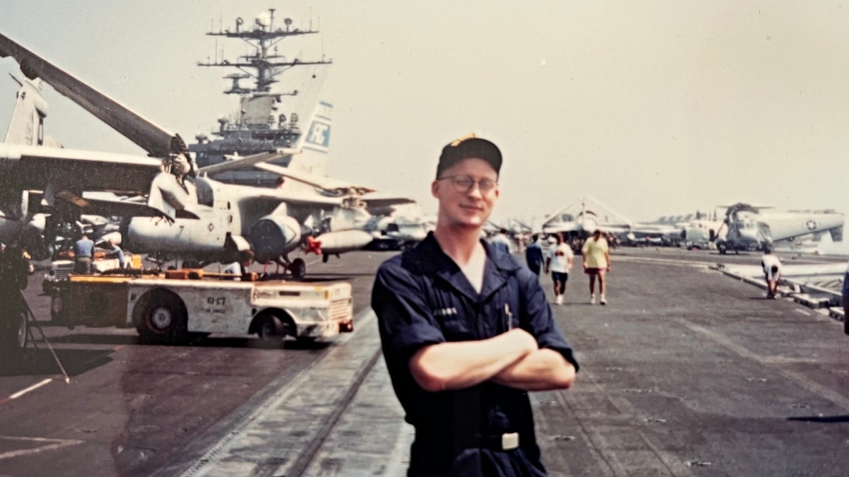 Philip Paxson poses in a Navy uniform with a Naval vessel in the background