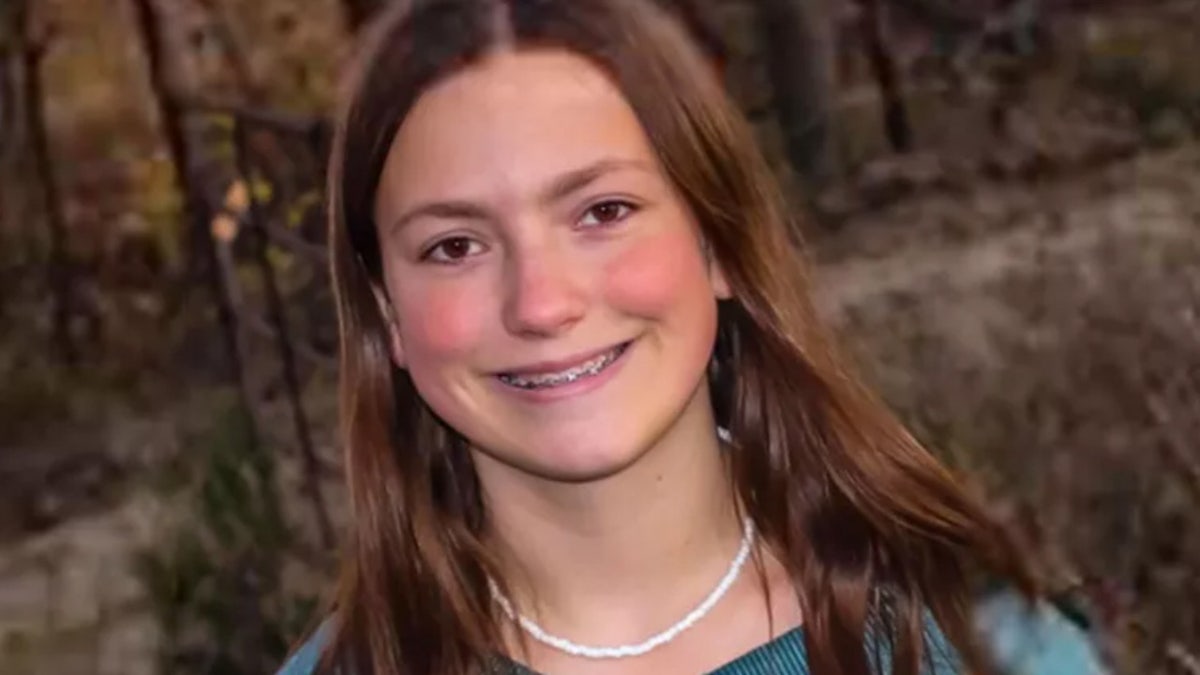 Young girl smiles in front of woods.