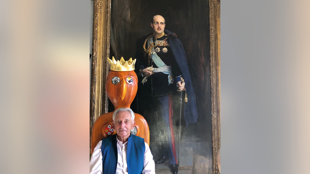 Prince Michael of Greece sitting in front of an art sculpture and royal portrait