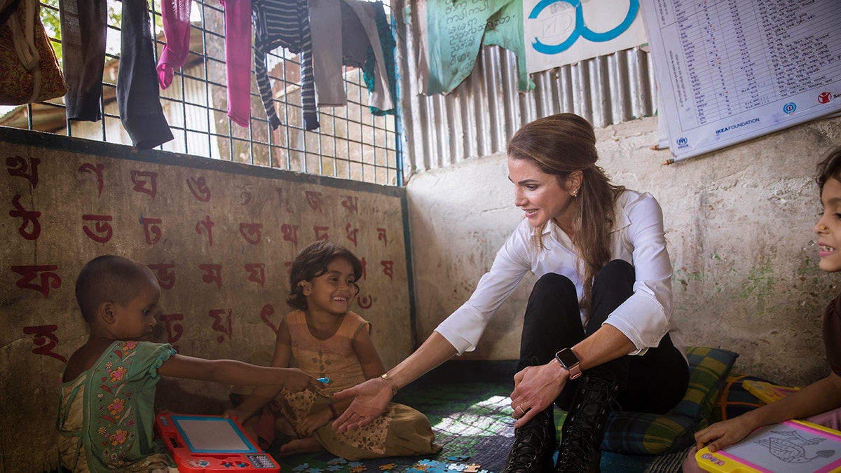 Queen Rania wearing a white blouse and black pants kneeling down in front of children