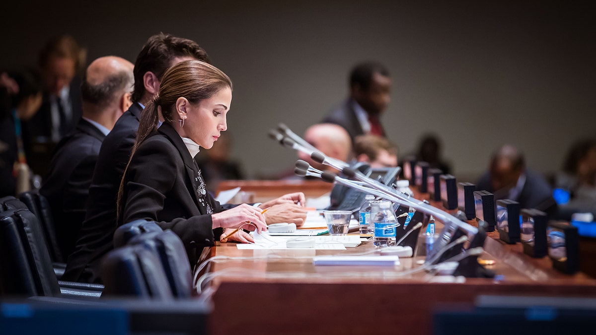 Queen Rania of Jordan wearing a navy suit and sitting on a desk looking down at a stack of papers