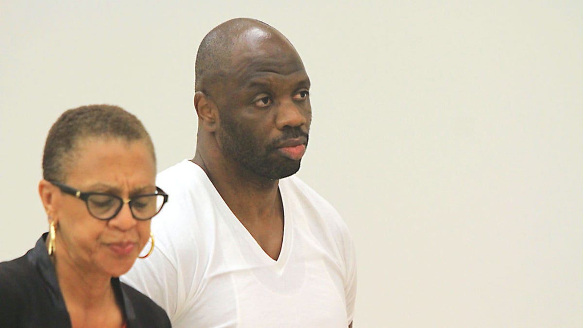 Somorie Moses in a white V-neck T-shirt stands next to his lawyer in court