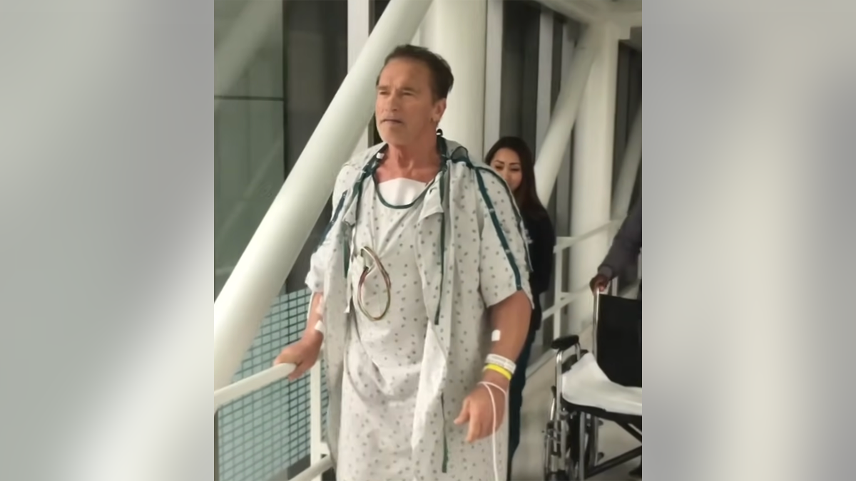 Arnold Schwarzenegger em uma bata de hospital andando pelo hospital