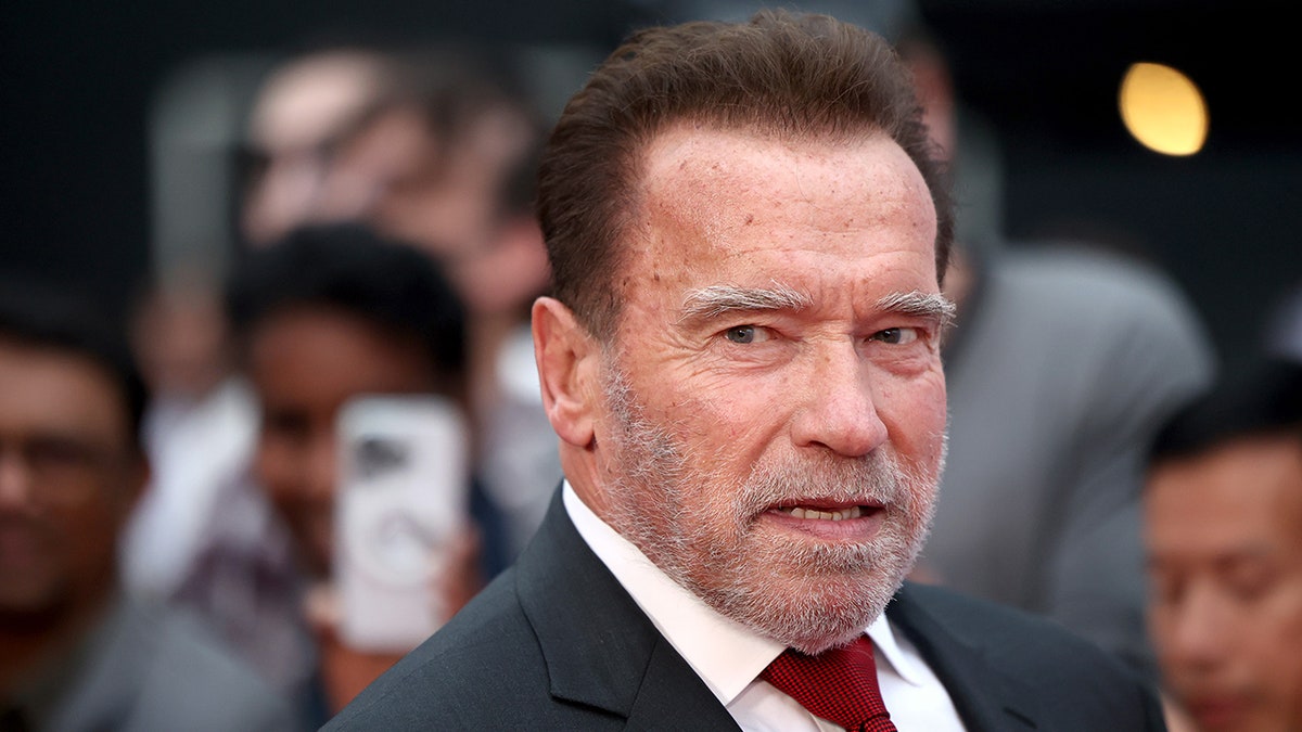Arnold Schwarzenegger parece sério no tapete em terno escuro e gravata vermelha