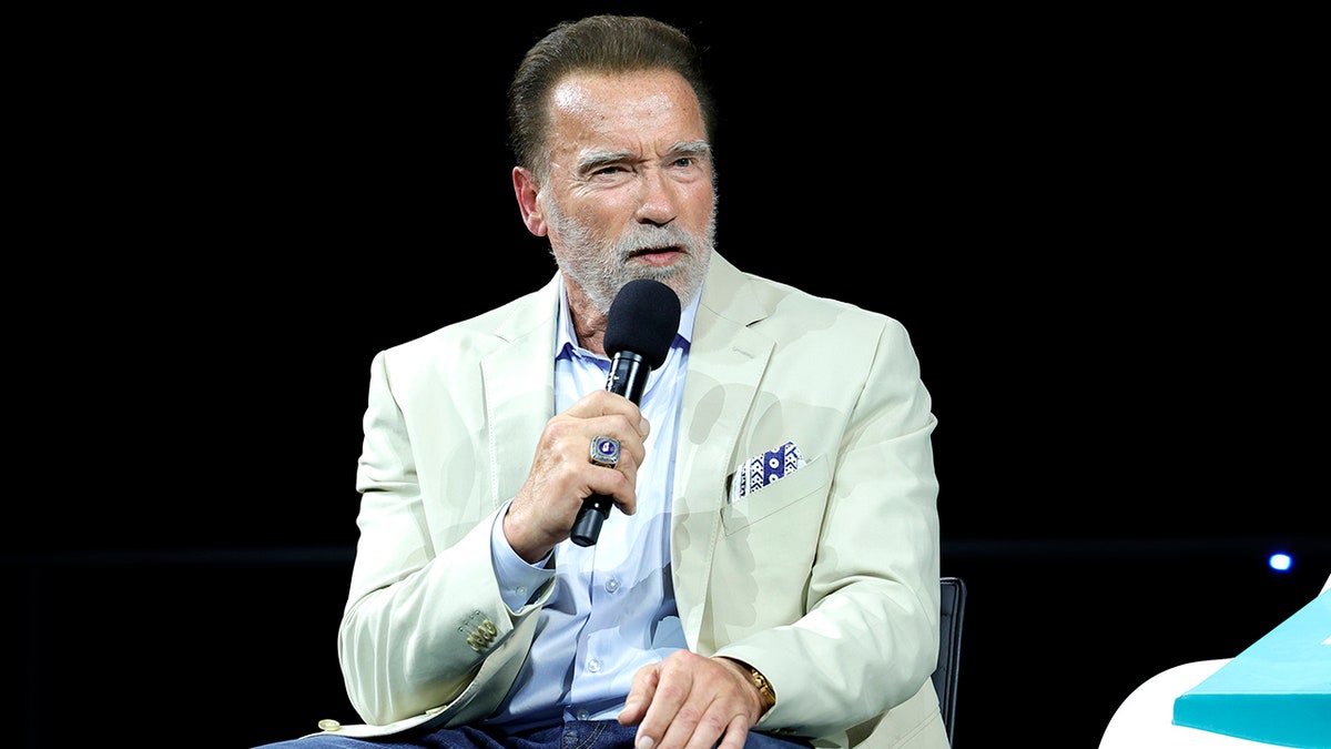 Arnold Schwarzenegger in a light blue shirt and off-white suit speaks into the microphone on stage