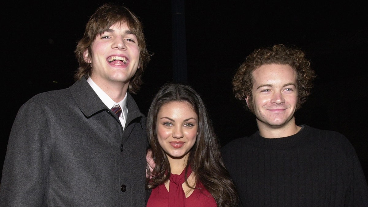 Ashton Kutcher smiles in a sweater and tie next to Mila Kunis in a red shirt next to Danny Masterson in 2000