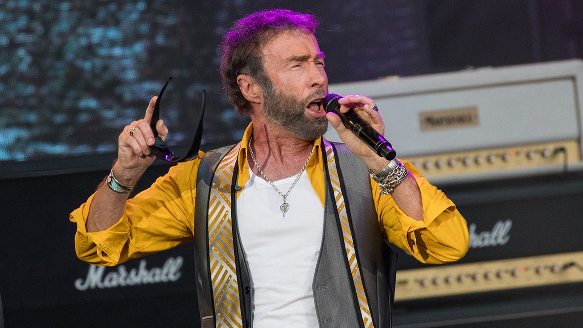 Paul Rodgers in a mustard yellow shirt and black vest with yellow tie sings on stage