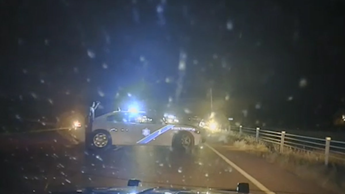 Arkansas Police Officer standing next to car looking out at white car that drove off side of highway
