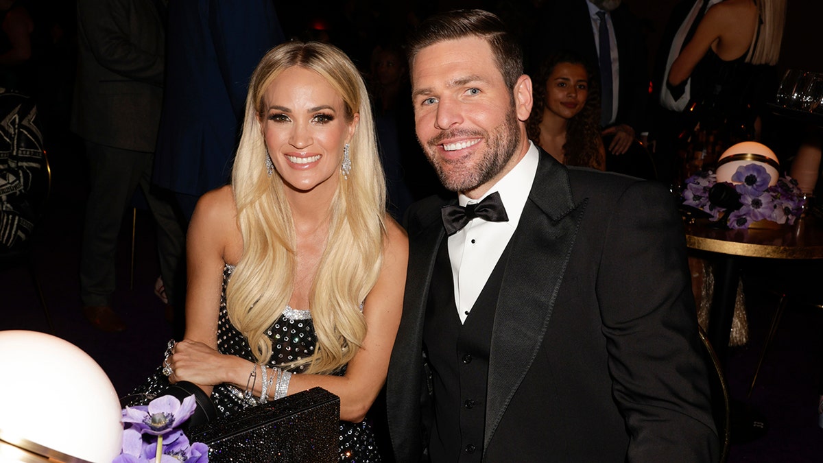Carrie Underwood with long blond hair sits next to her husband Mike Fisher in a tuxedo at a table at the Grammy Awards