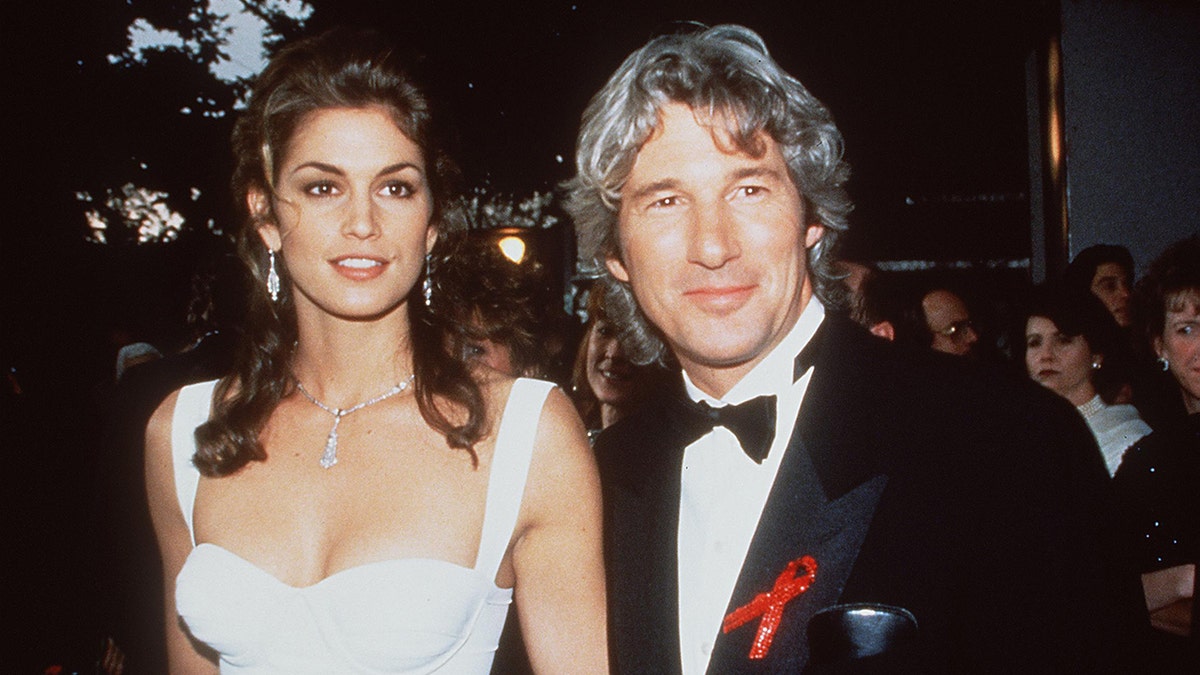 Cindy Crawford in a white gown and dangly earrings poses for a photo with a grinning Richard Gere in a classic tuxedo with a red ribbon
