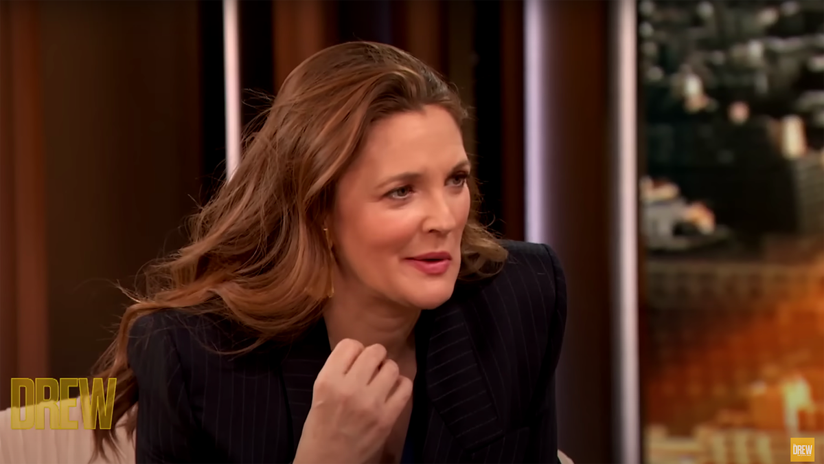 Drew Barrymore in a black blazer leaning over in an interview on 