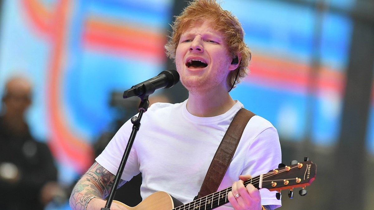 Ed Sheeran in a white shirt closes his eyes as he performs in New York City with a guitar around his shoulder