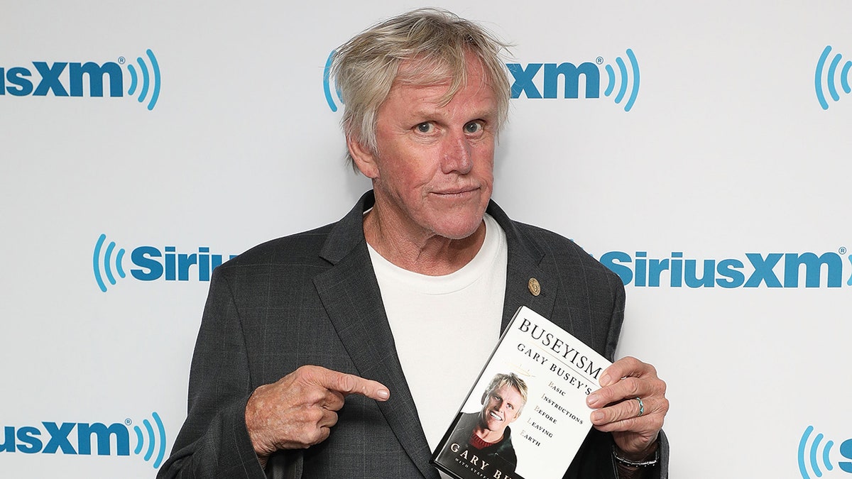 Gary Busey points to his book that he's holding up and smirks at the camera