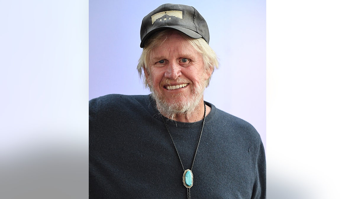 Gary Busey in a black shirt and necklace with a turquoise stone wears a black hat slightly atop his head