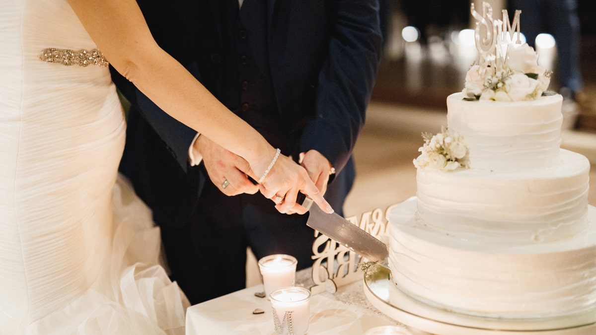 A bride and groom cut a three-tiered cake decorated in white frosting together at a wedding ceremony.