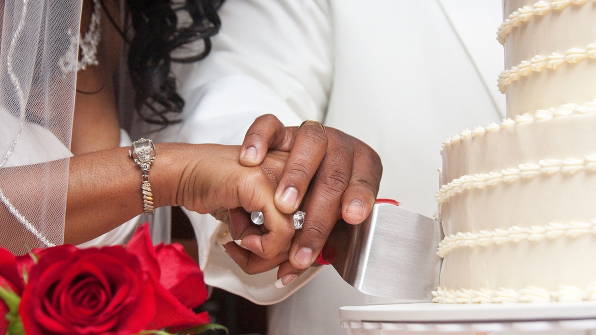A bride and groom cut their wedding cake together with red roses nearby.