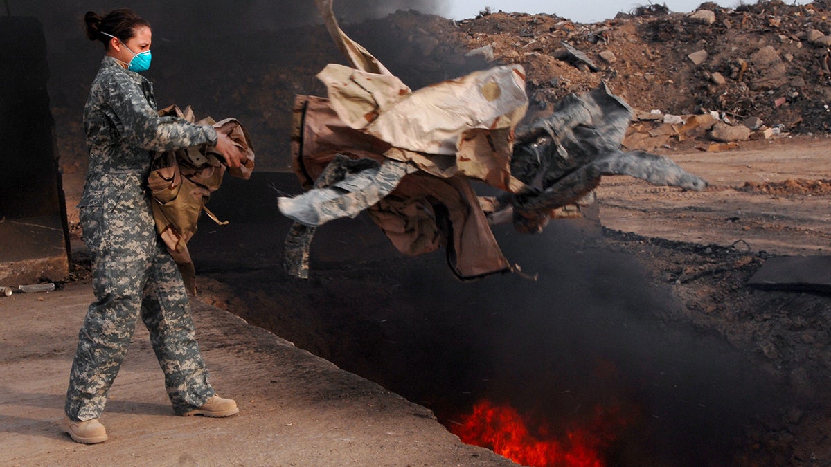 A female airman wearing a mask throws uniforms into a burn pit
