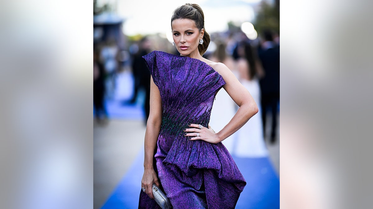 Kate Beckinsale in a dark purple gown puts her hand on her hip as she attends the amfAR Gala in Cannes