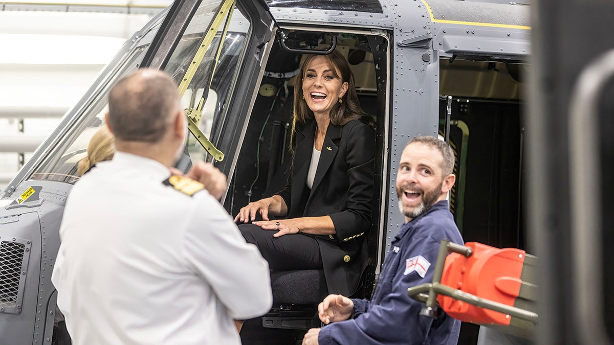 Kate Middleton in a black suit and pant set laughs as she sits in a helicopter