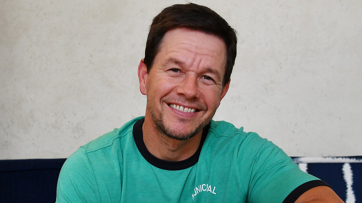 Mark Wahlberg in a teal t-shirt smiles for a picture why showcasing his tequila