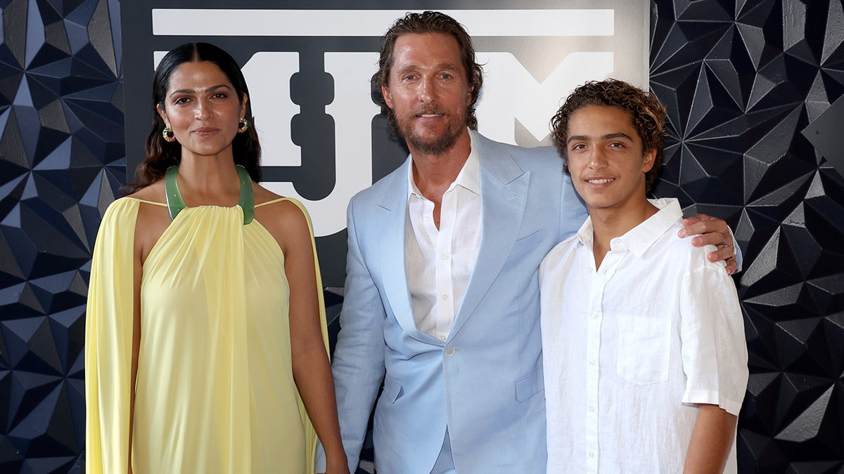Camila Alves in a yellow gown with green halter straps smiles next to Matthew McConaughey in a blue suit and pants standing next to their son Levi in a white button down on the carpet in Texas
