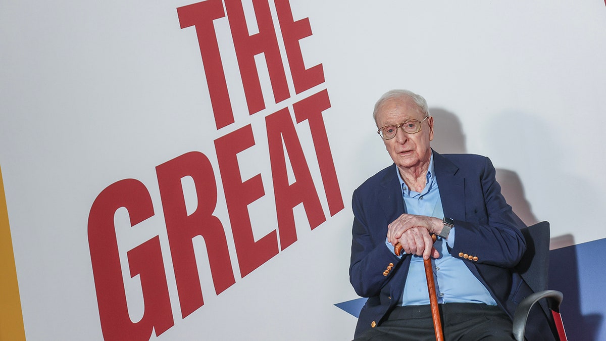Michael Caine sitting in front of a "Great Escaper" poster