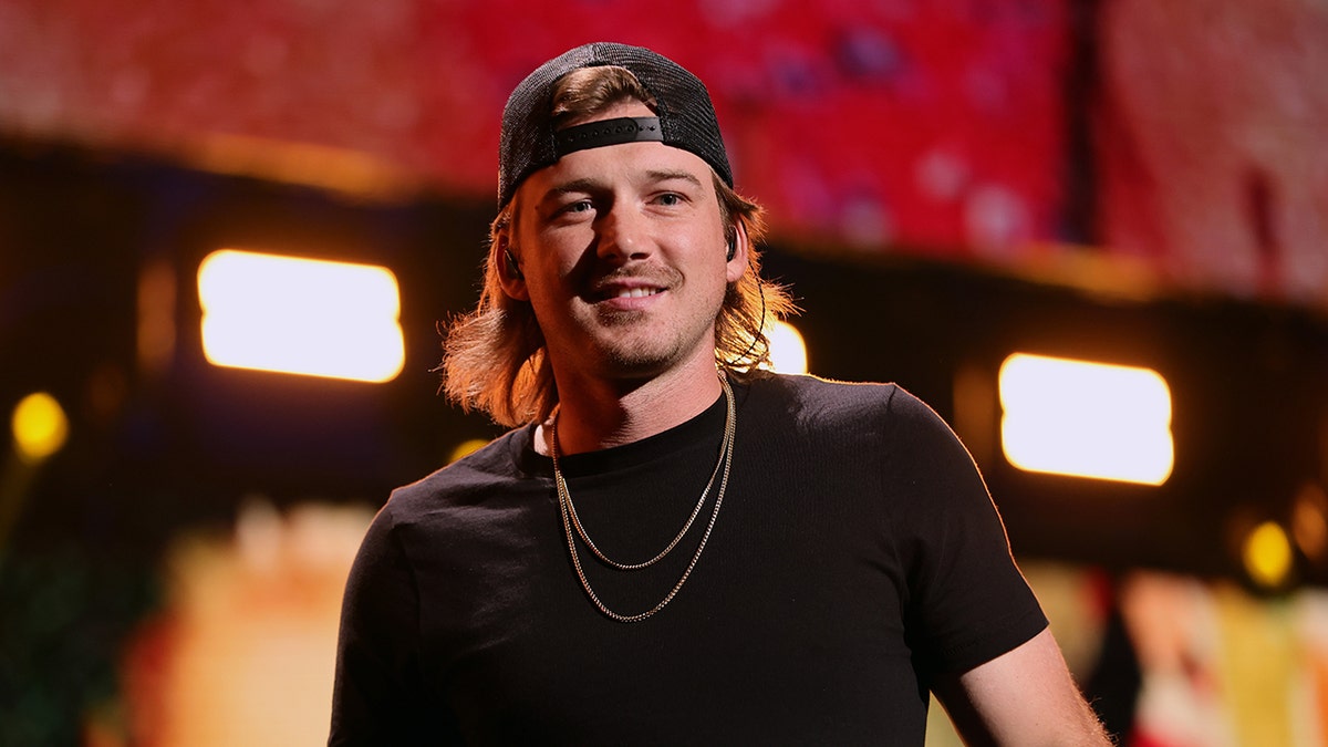 Morgan Wallen performing in a black shirt and chains and backwards hat