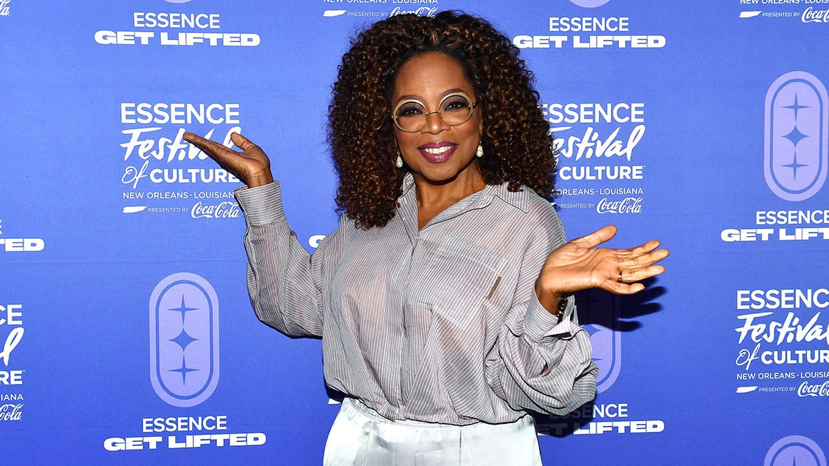 Oprah Winfrey with her hands out on either side in the air posing on the carpet in a grey top and white pants